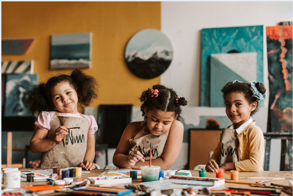 Three children sitting at a table with art supplies in a classroom setting.
